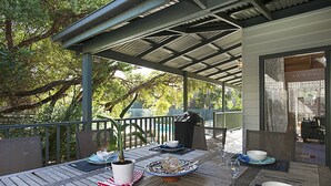 Un espace extérieur aménagé pour les repas, avec un patio couvert, une table en bois, des chaises, des plantes en pot et une vue sur les arbres et le ciel environnants.