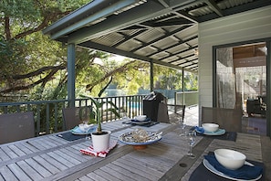 An outdoor dining area with a covered patio, wooden table, chairs, potted plants, and a view of the surrounding trees and sky. - Poolside Sorrento Pet Friendly (SORRENTO)