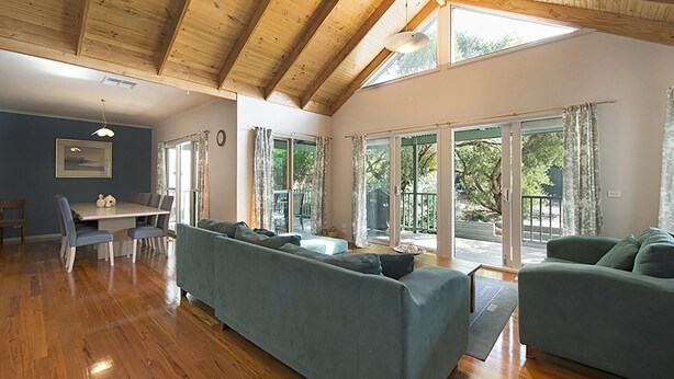 A spacious living area with a wooden ceiling, a dining table with chairs, a sofa, and a large sliding glass door leading to an outdoor area. - Poolside Sorrento (SORRENTO)