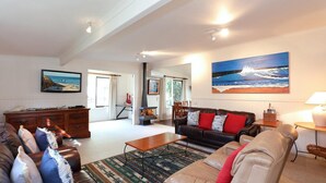 A spacious living room with a fireplace, a large painting of a coastal scene, and a dining area. - Back Beach Hideaway - Family friendly beach house (BLAIRGOWRIE)