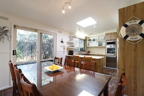 A modern kitchen with a dining area, featuring a large wooden table, chairs, and a view through the glass doors. - Back Beach Hideaway Blairgowrie (BLAIRGOWRIE)