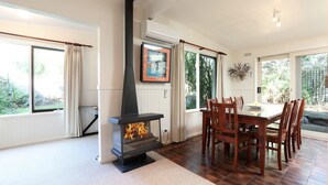 A dining area with a wood-burning stove, a dining table with chairs, and large windows with curtains. - Back Beach Hideaway - Family friendly beach house (BLAIRGOWRIE)
