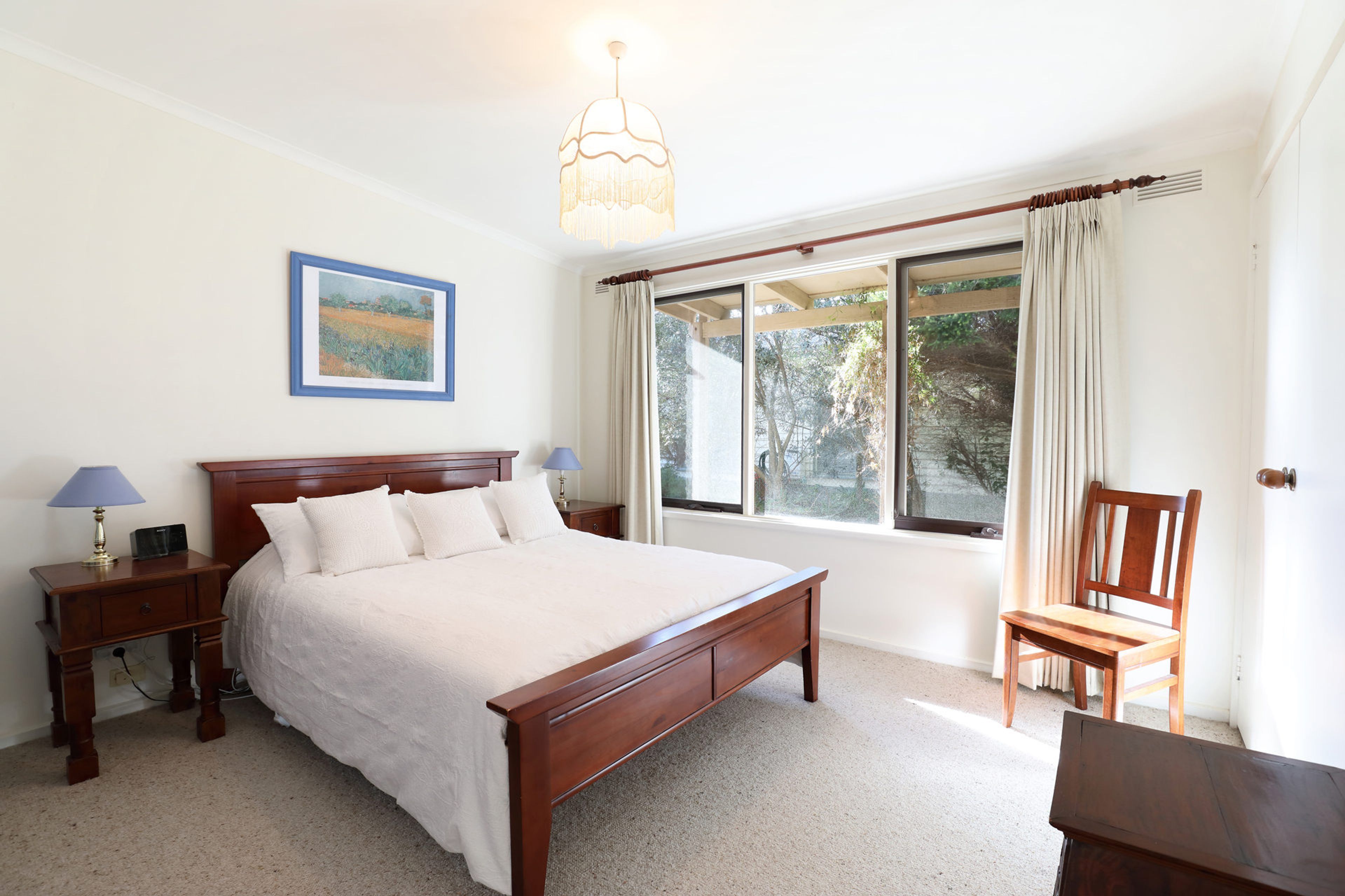 A bedroom with a large bed, a bedside table with a lamp, a chair, a window with curtains, and a painting on the wall.