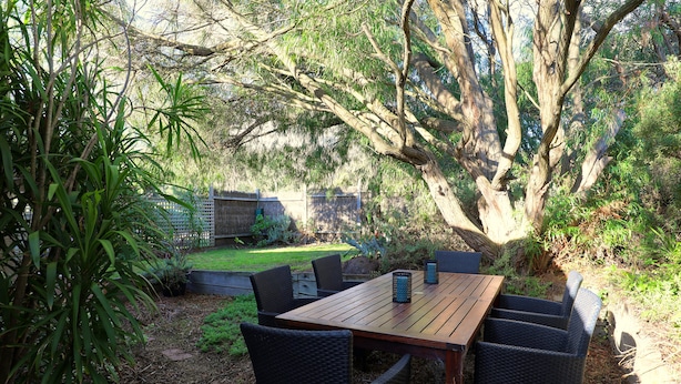 Een terras buiten met een houten tafel en rieten stoelen, omgeven door bomen en een gazon.