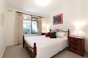 A bedroom with a bed, two bedside tables with lamps, a window with curtains, and a wall-mounted picture. - Back Beach Hideaway Blairgowrie (BLAIRGOWRIE)