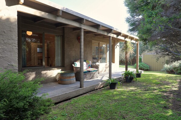A covered outdoor area with a wooden deck, a seating area with cushions, and potted plants. - Back Beach Hideaway Blairgowrie (BLAIRGOWRIE)