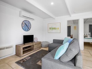 A modern living room with a grey sofa, a flat-screen TV on a wooden stand, a large wall clock, and a framed map. - Quarterdeck Portsea Coastal Pet Friendly Escape (PORTSEA)