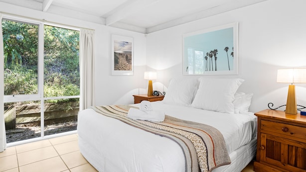 A hotel room with a bed, two bedside tables, a window with a view of greenery, and a painting on the wall.