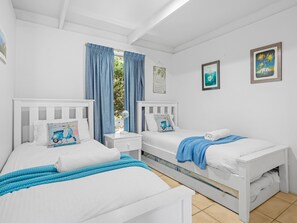 Una habitación con dos camas blancas, una ventana con cortinas azules y una pared con dos cuadros enmarcados.
