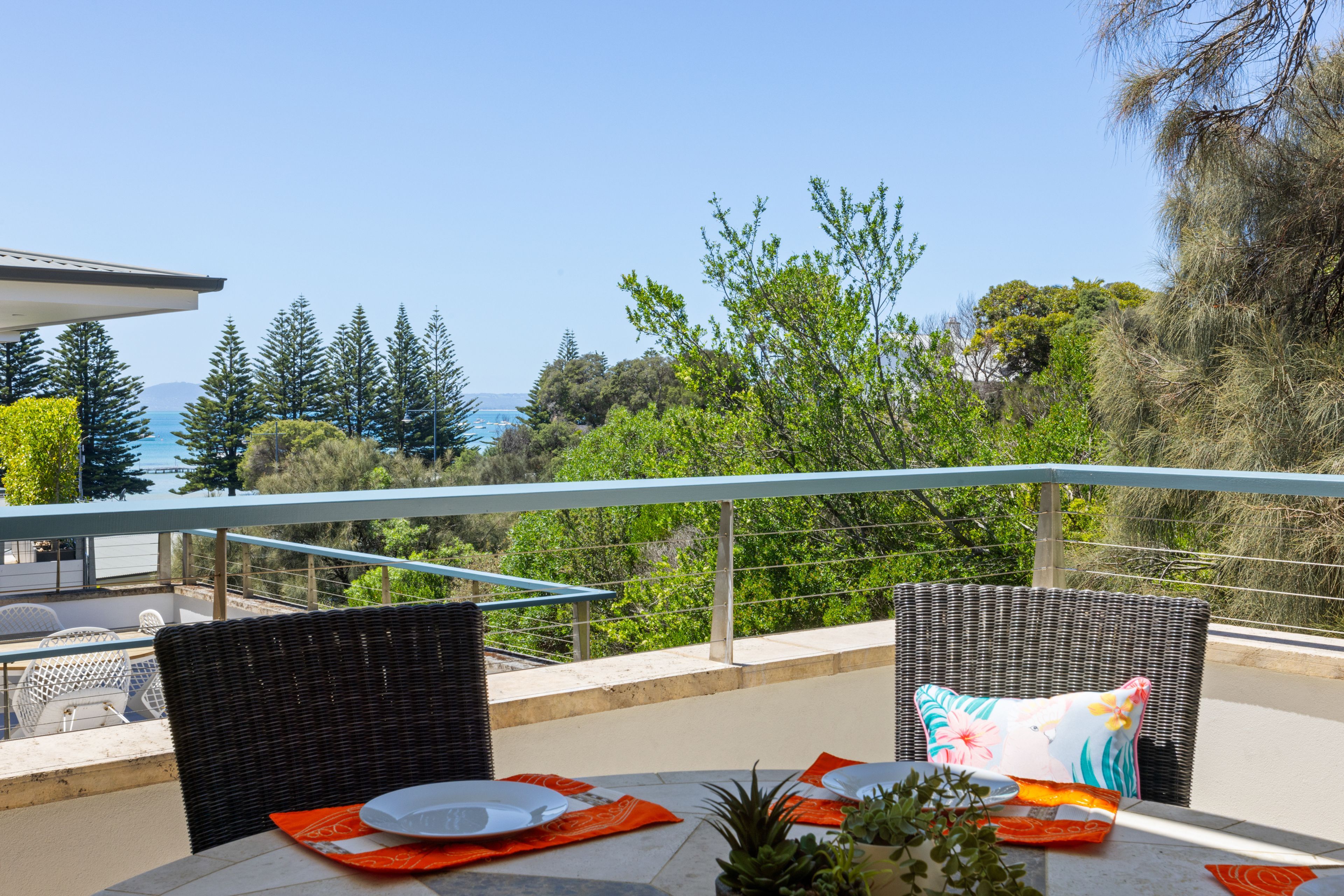 An outdoor dining area with wicker furniture, a table set with plates, and a view of the sea and trees.