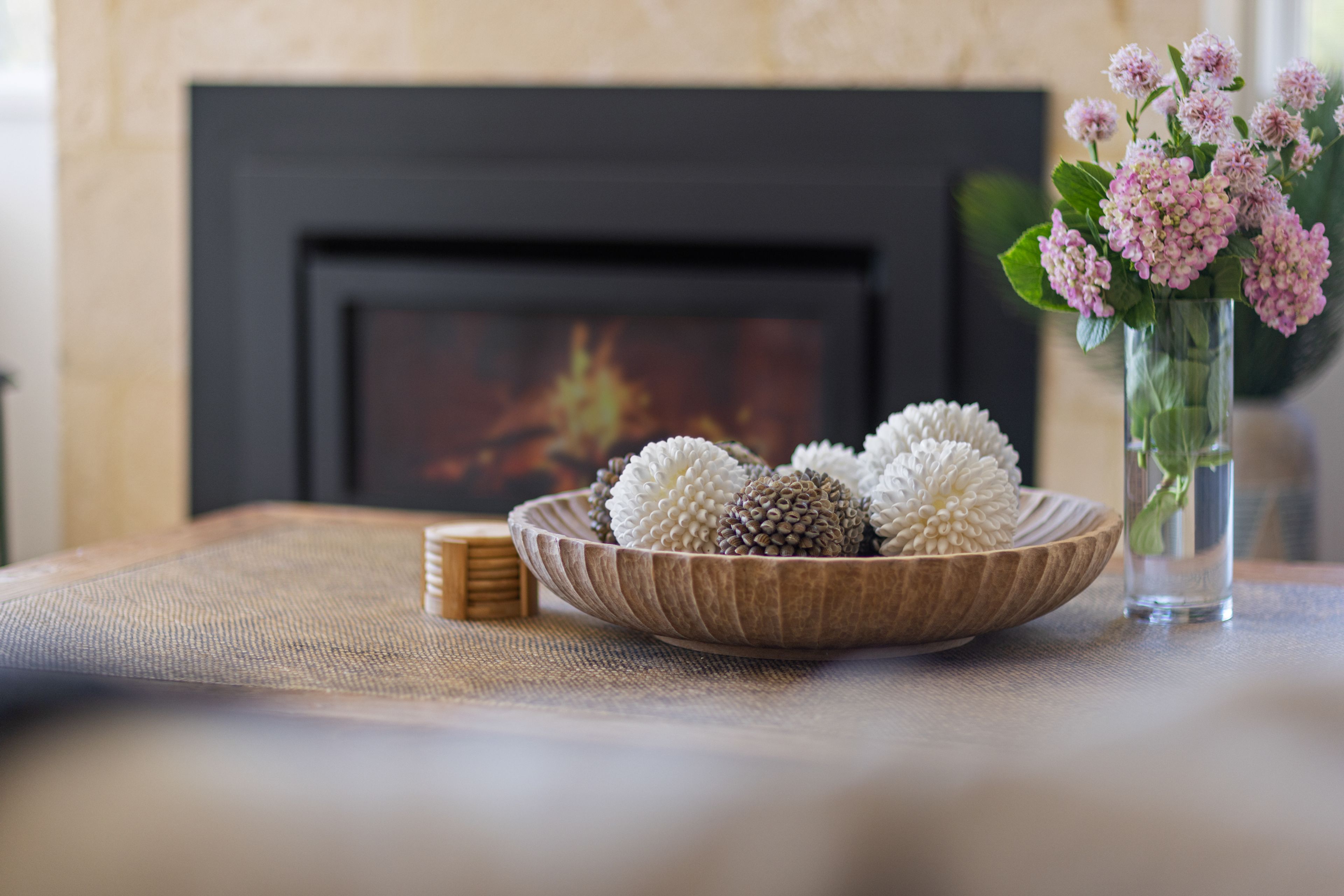 A wooden bowl with decorative items on a table, a vase with flowers, and a fireplace in the background.