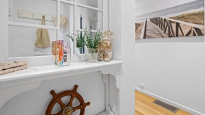 Une pièce avec une fenêtre, un gouvernail de bateau en bois, une étagère remplie de livres et d’objets décoratifs, et un tableau représentant une scène de plage accroché au mur.