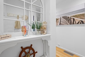 Una habitación con ventana, una rueda de barco de madera, una estantería con libros y objetos decorativos, y una pintura de una escena de playa en la pared.