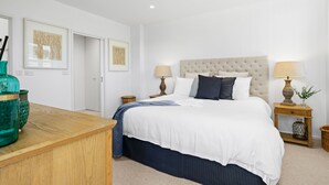 Une chambre à coucher avec un grand lit, deux tables de chevet avec des lampes, une commode en bois et deux œuvres d’art encadrées accrochées au mur.