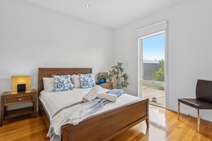 A bedroom with a wooden bed, a nightstand, a lamp, a potted plant, and a view of the outdoors. - Sorrento City Style Near Ocean Beach (SORRENTO)