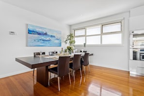 Uma sala de jantar com mesa de madeira posta para quatro pessoas, um quadro acima da mesa e uma janela com persianas.