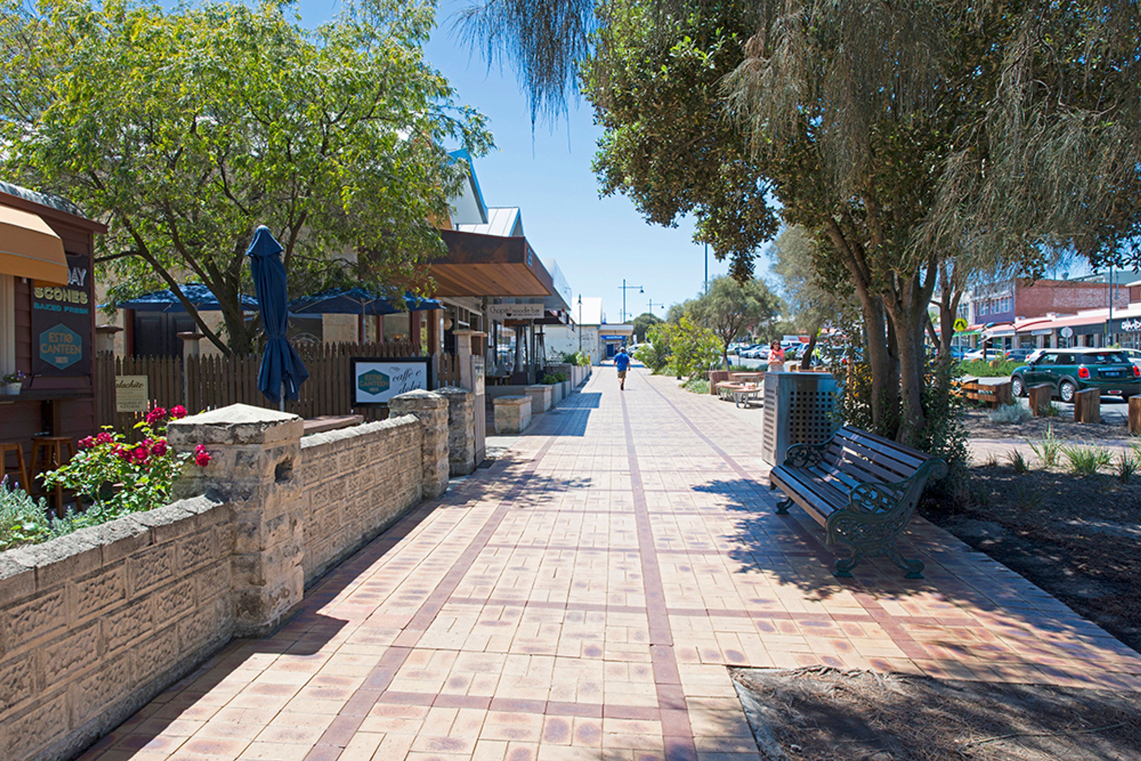 A paved walkway with a bench, a stone wall, and a row of buildings with awnings.