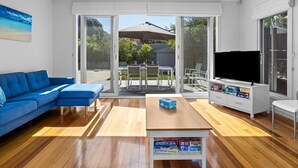A modern living room with a blue sofa, a wooden coffee table with board games, a TV, and a sliding glass door leading to an outdoor patio. - Bluetopia Spacious Rye Retreat with Pool and BBQ (RYE)