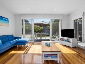 Un salon moderne avec un canapé bleu, une table basse en bois sur laquelle sont posés des jeux de société, un téléviseur et une porte-fenêtre coulissante donnant sur une terrasse extérieure.