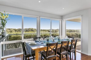 A dining area with a table set for six, large windows offering a view of the countryside, and a potted plant on the left. - Dunescape Spacious Rye Retreat with Private Pool (RYE)