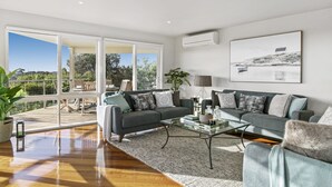 A spacious living room with a large window, a grey sofa set, a coffee table, and a painting on the wall. - Dunescape Spacious Rye Retreat with Private Pool (RYE)