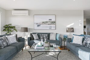 A modern living room with a grey sofa set, a coffee table, and a painting of a coastal scene. - Dunescape Spacious Rye Retreat with Private Pool (RYE)
