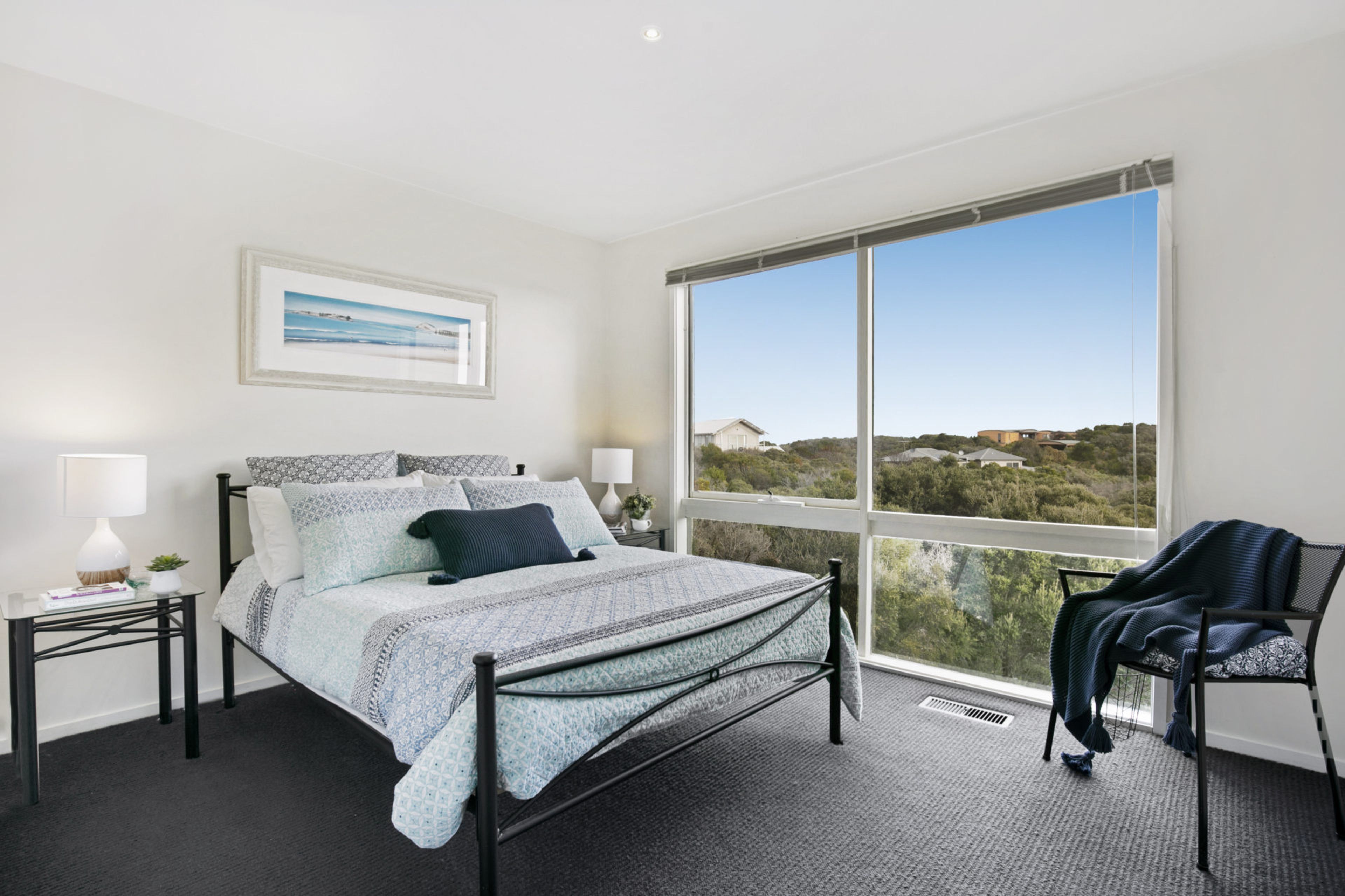 A bedroom with a large window, a bed with patterned bedding, a bedside table with a lamp, and a chair.