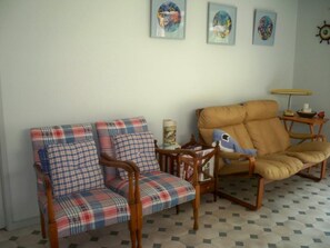 Ein Wohnzimmer mit zwei Holzstühlen, einer mit Kissen in Karomuster und der andere mit einfarbigem Kissen, einem Holztisch mit Lampe sowie drei gerahmten Kunstwerken an der Wand.