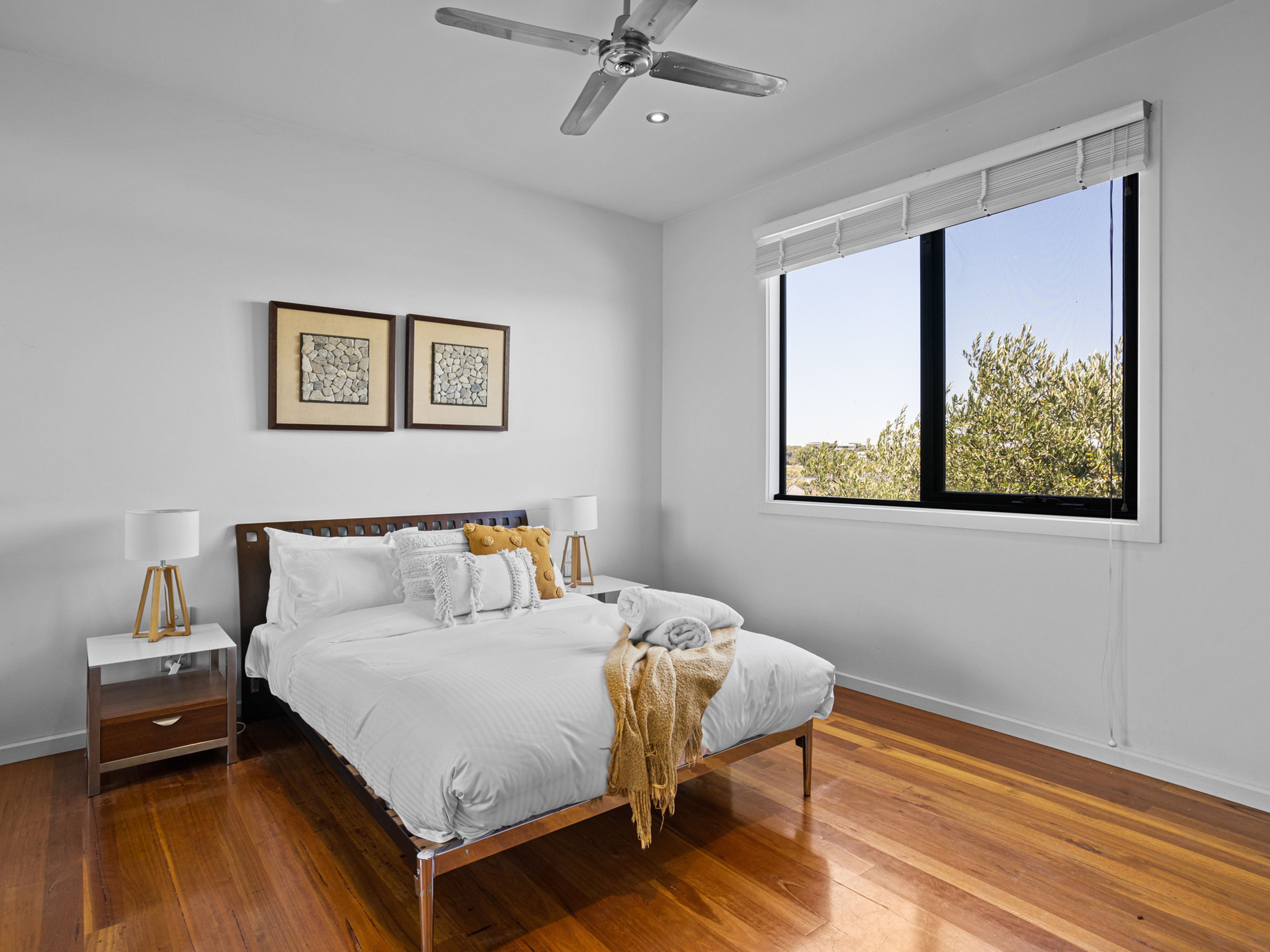 A bedroom with a bed, bedside tables, a ceiling fan, framed artwork, and a large window with a view.