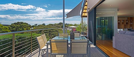 Un balcon avec une table et des chaises en verre, une terrasse en bois et une vue sur le ciel et les arbres.