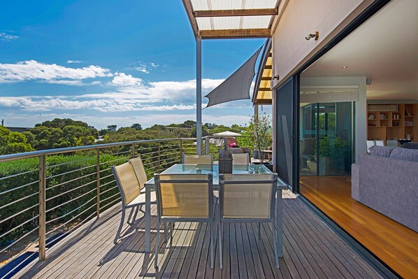 A balcony with a glass table and chairs, a wooden deck, and a view of the sky and trees.