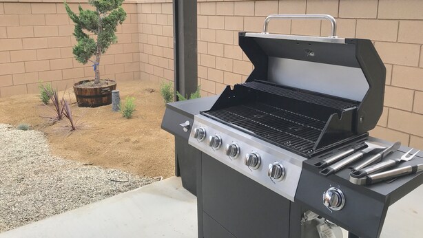 Un jardin avec un barbecue noir doté de quatre brûleurs et d’une étagère latérale où sont rangés les ustensiles de cuisson.