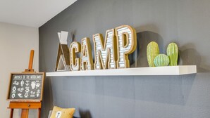 A room with a grey wall, a white shelf displaying cacti and the word "CAMP", a wooden easel with a chalkboard, and a grey sofa with a yellow pillow. - 1000 MBPS Fast Wifi★Model Home★Luxury Family Retreat 5B/4b (Jurupa Valley)
