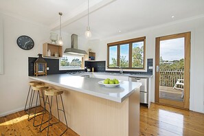 A modern kitchen with a central island, bar stools, a clock, and a view through large windows. - Treetops Coastal Family Retreat in Sorrento (SORRENTO)