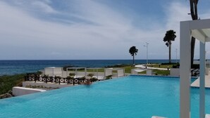 A swimming pool with a view of the ocean, surrounded by white structures and palm trees.