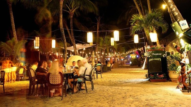 An outdoor dining area with tables and chairs, palm trees, and string lights.