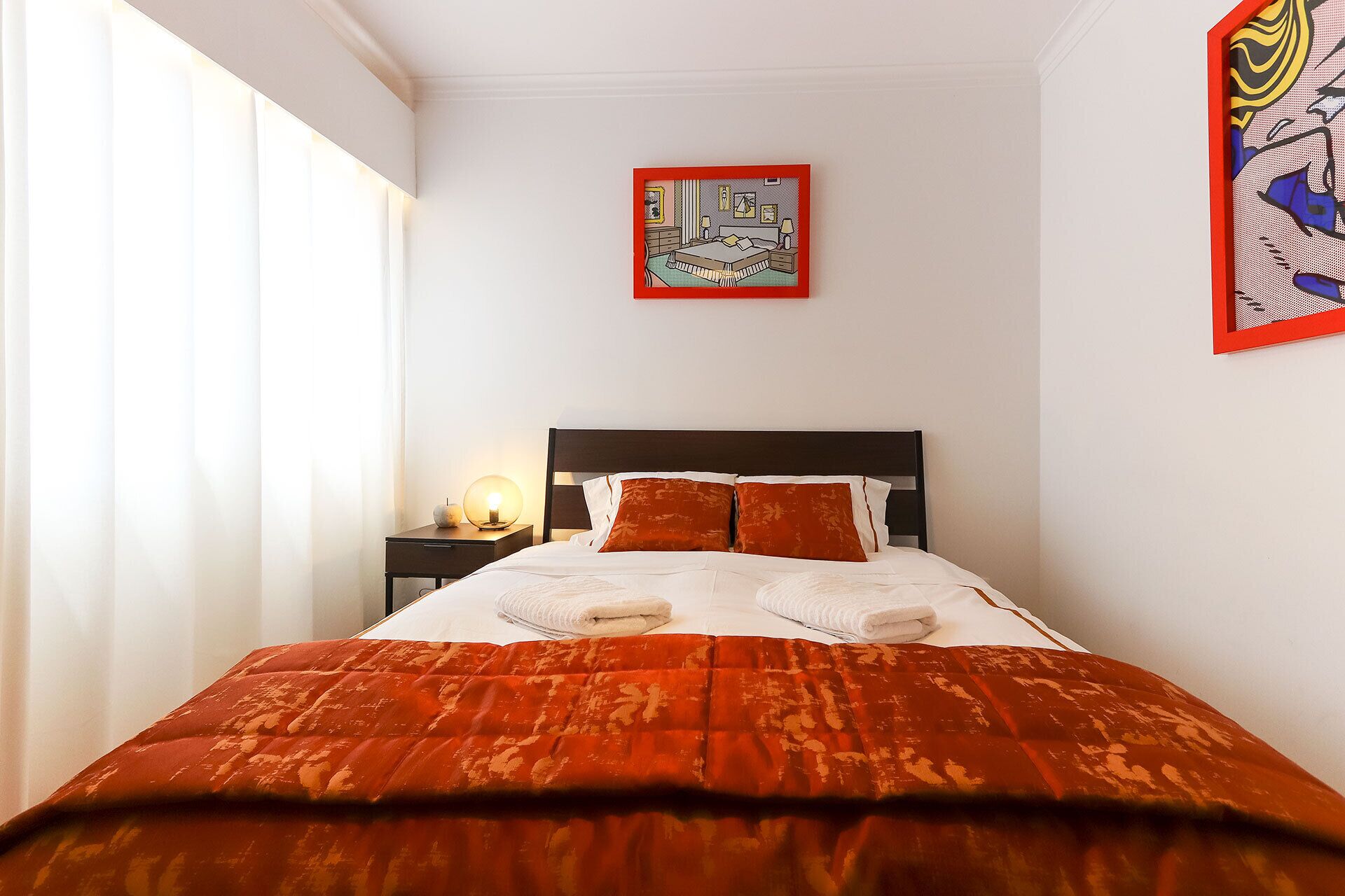 A neatly made bed with a red and white patterned bedspread, a wooden headboard, and two pillows. There is a bedside table with a lamp and a small decorative item. A framed artwork hangs above the bed on the wall.
