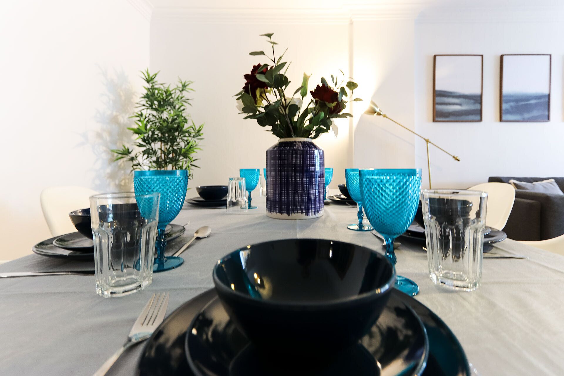 A dining table set with blue glassware, a black bowl, and a vase with flowers.