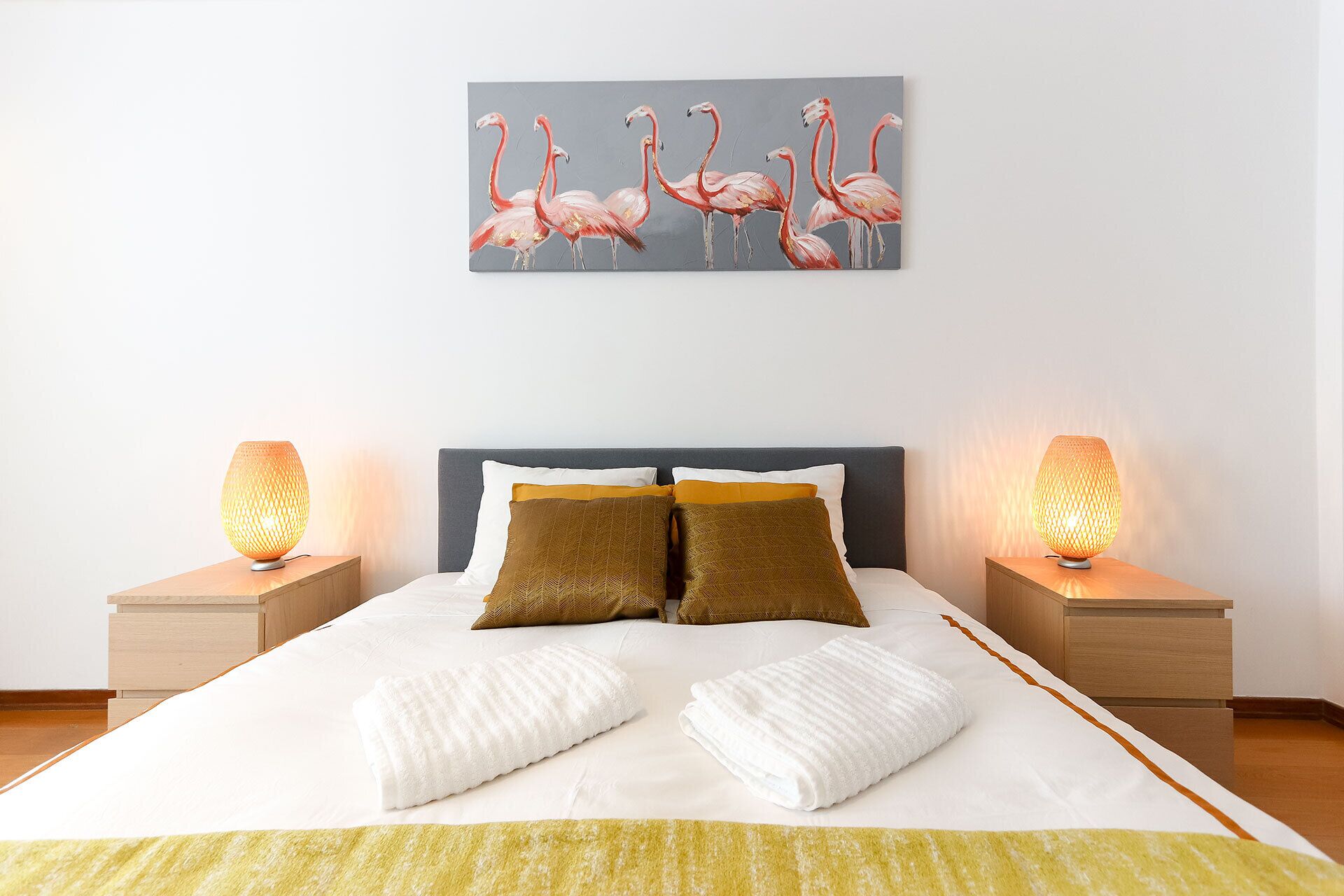 A neatly made bed with two pillows, a bedside table with a lamp, and a wall-mounted painting of flamingos.