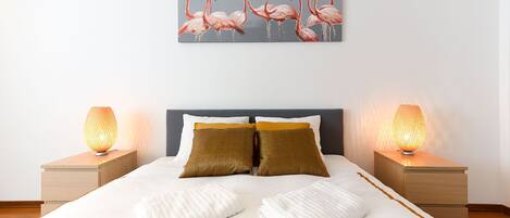 A neatly made bed with two pillows, a bedside table with a lamp, and a wall-mounted painting of flamingos.