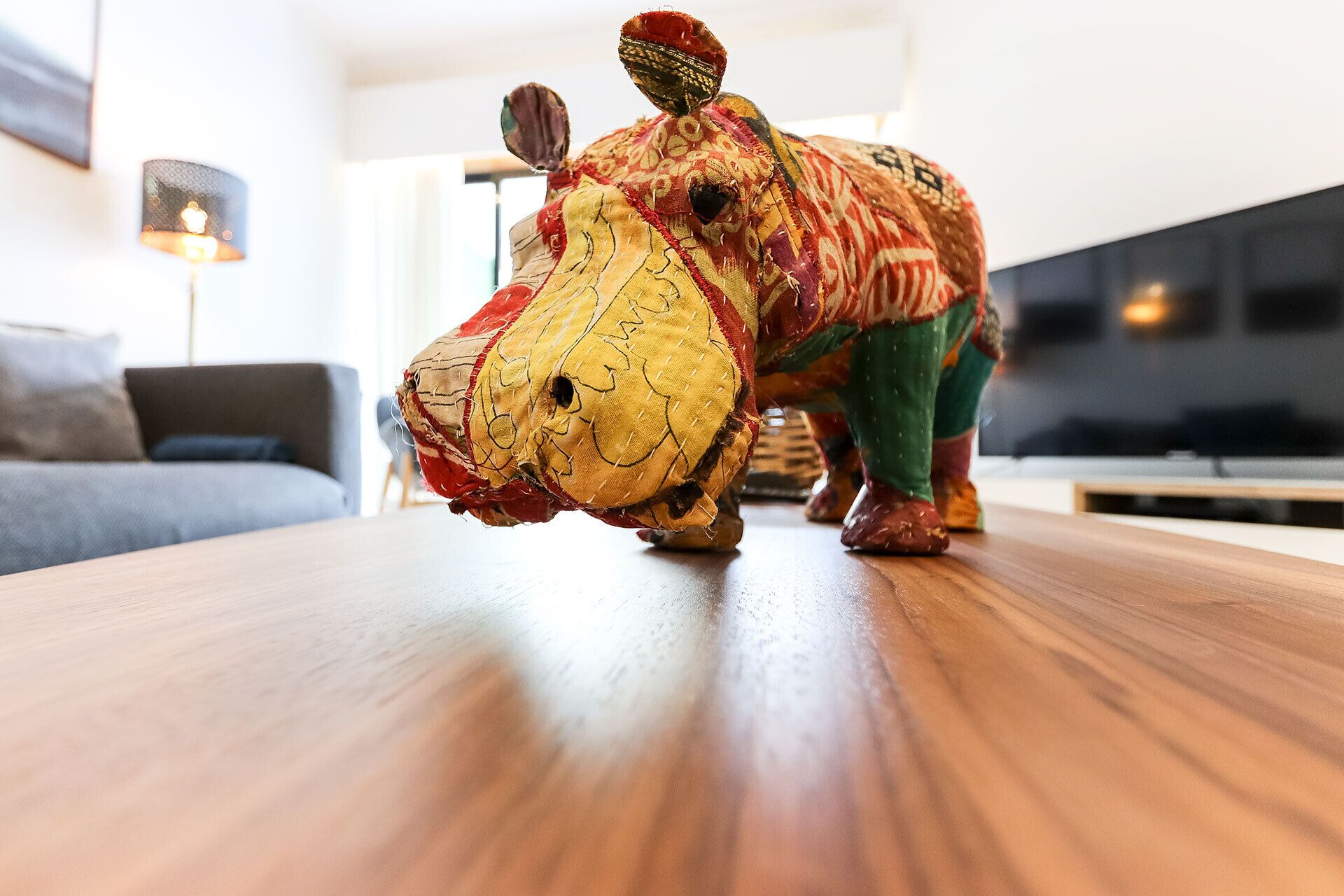 A plush toy on a wooden table in a living room with a sofa and a lamp in the background.