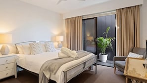 Una habitación de hotel moderna con cama, mesitas de noche, una silla, una planta en maceta y un ventanal corredero al balcón.