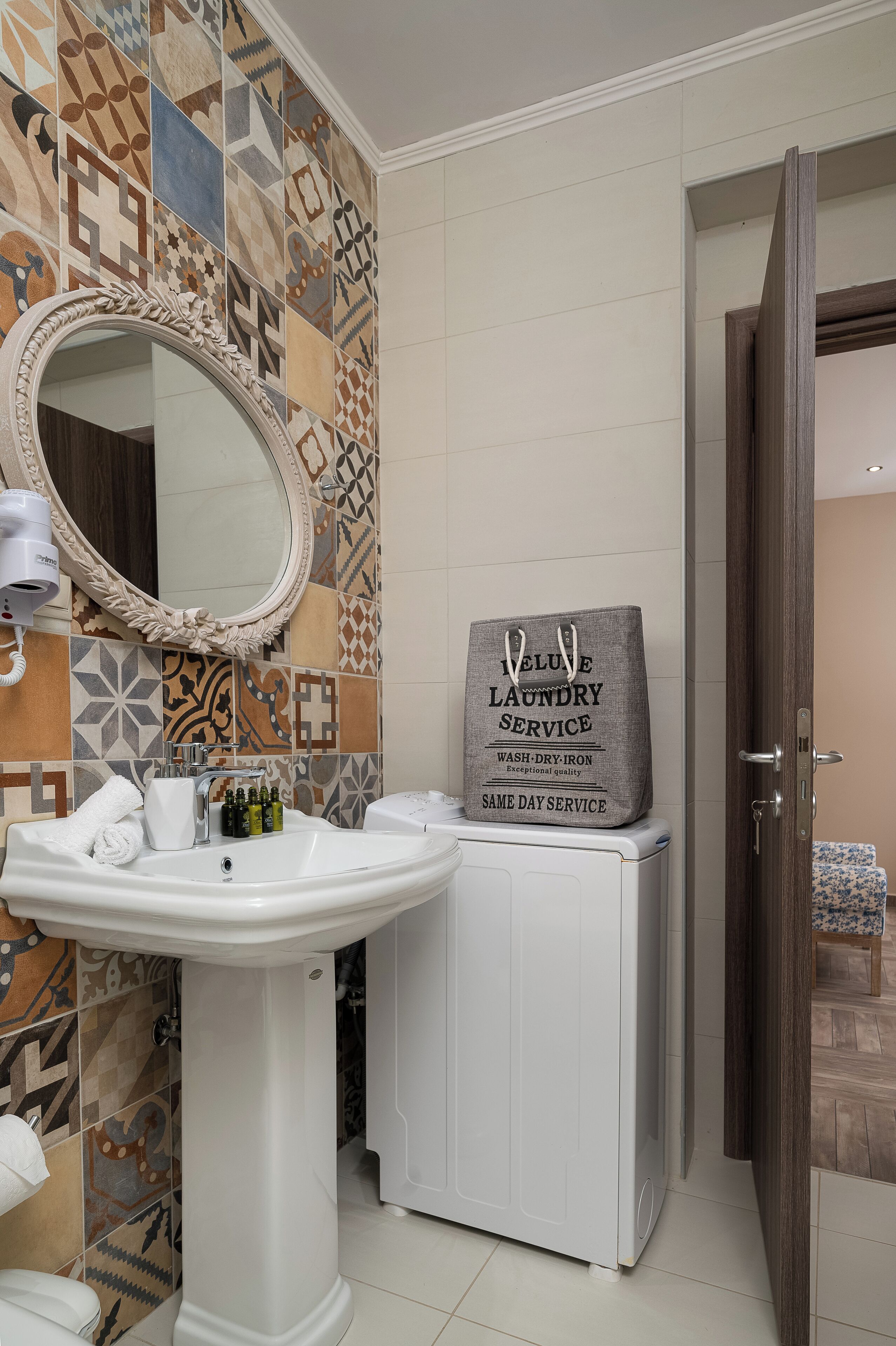 Une salle de bain avec un mur carrelé, un lavabo, un miroir et un sac à linge posé sur un meuble.