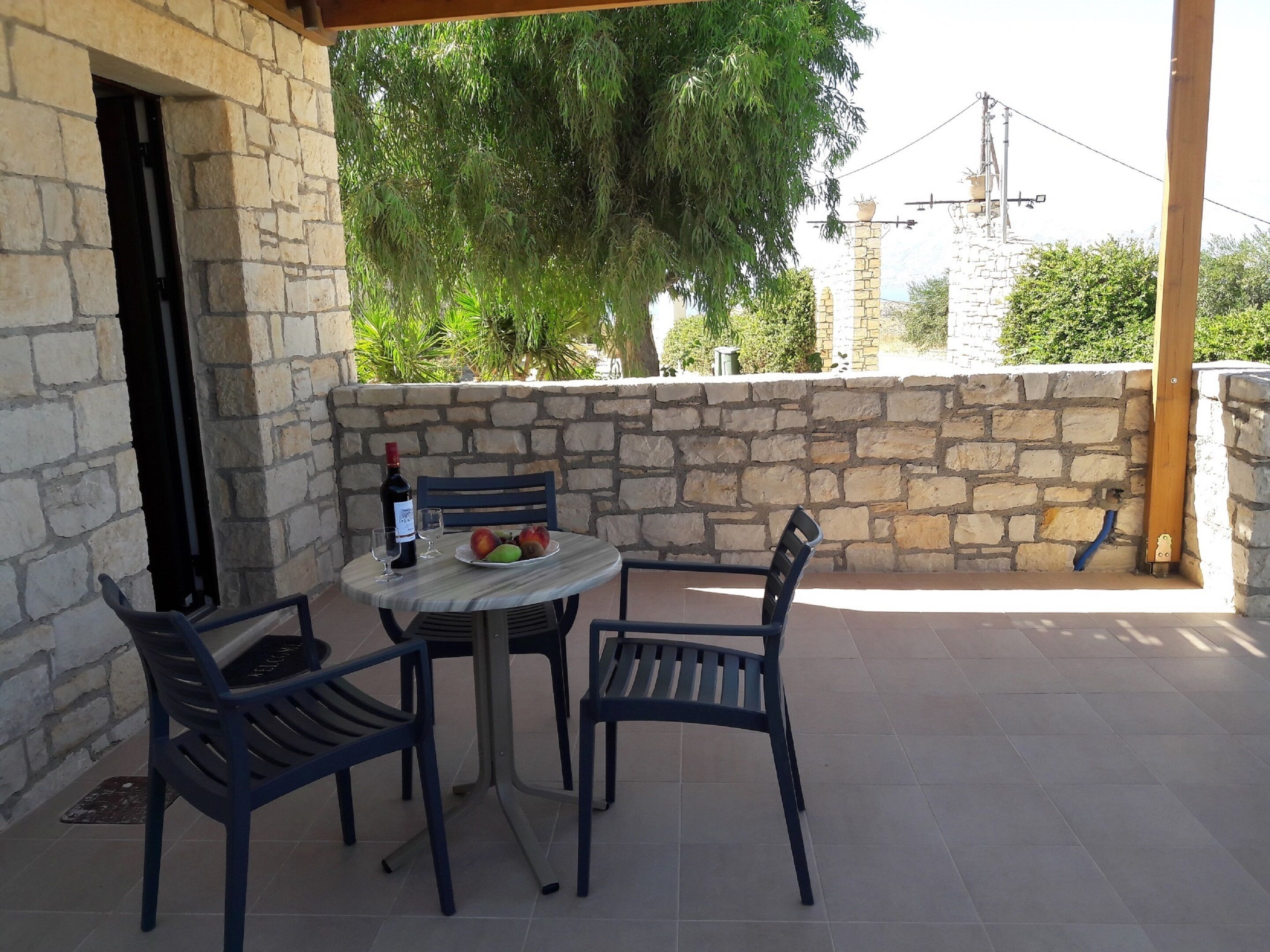 A stone-walled outdoor patio with a table and chairs, a bottle of wine, and a plate of fruit.