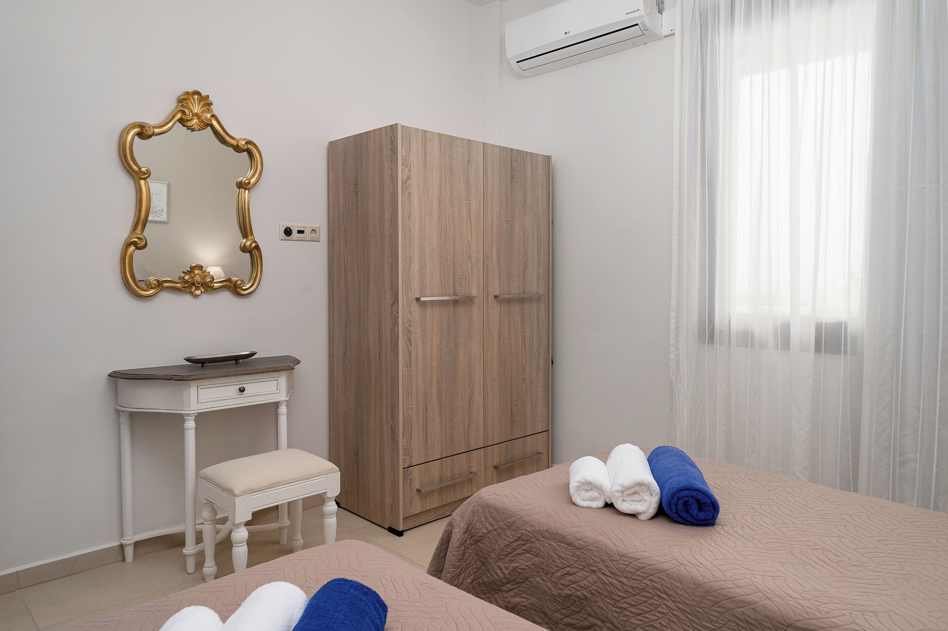 Habitación de hotel con un armario de madera, un pequeño escritorio blanco con una silla, una cama con dos toallas enrolladas y un espejo grande con un marco decorado.