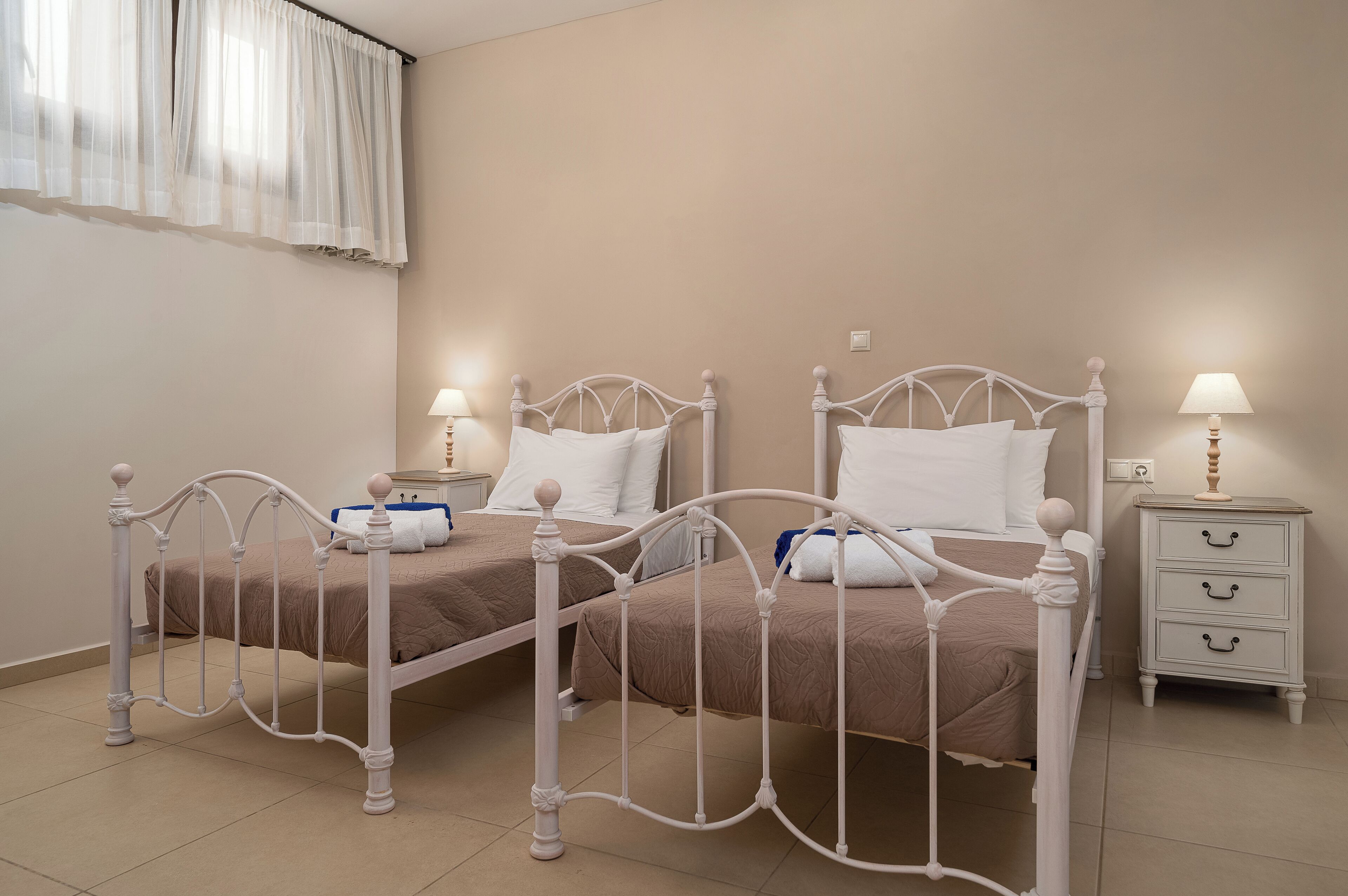 Un lit double avec une tête de lit et un pied de lit en métal blanc, deux tables de chevet avec des lampes et une fenêtre avec des rideaux légers.
