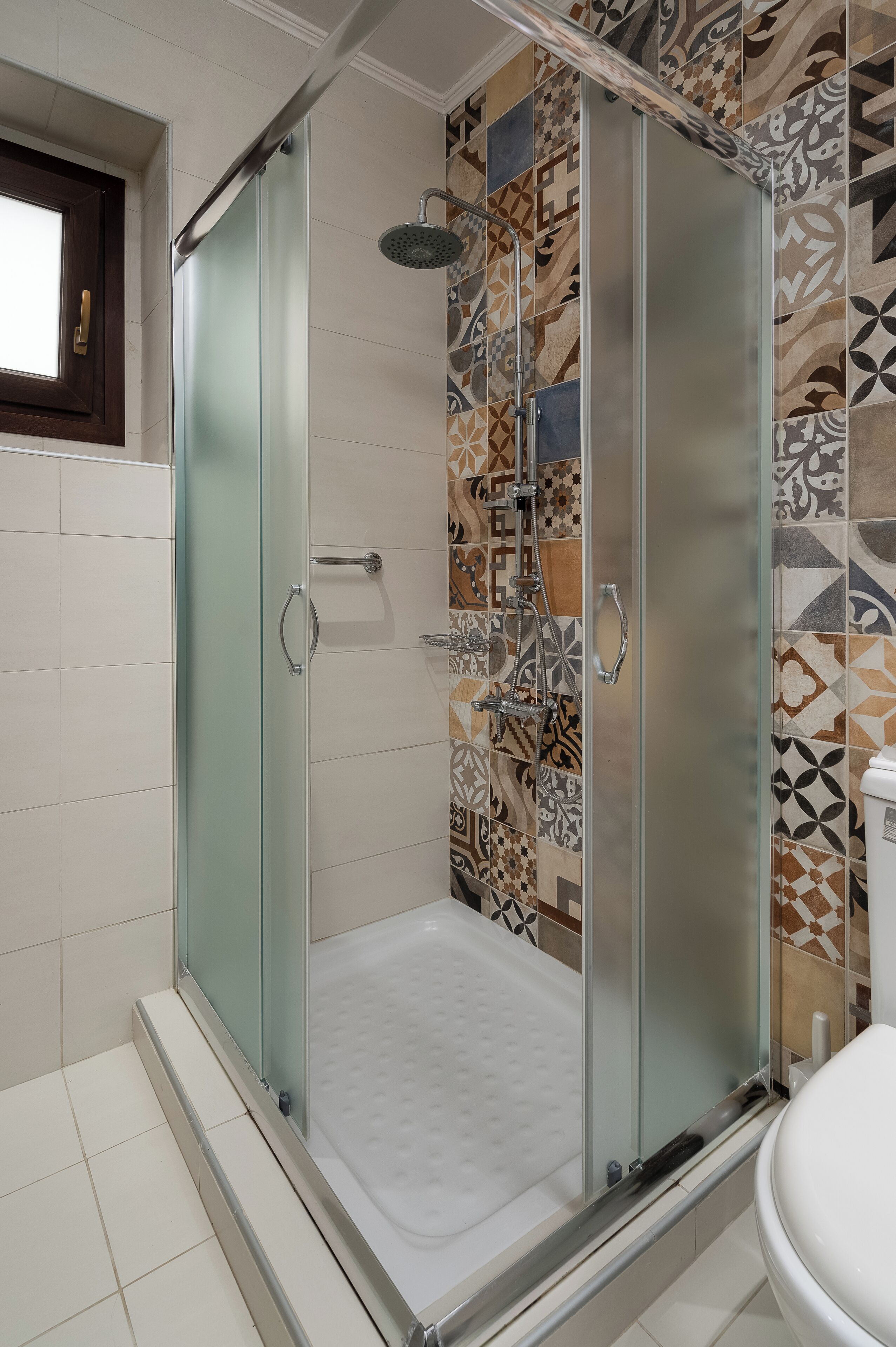 A glass-enclosed shower with a patterned tile wall, a white toilet, and a window with a wooden frame.