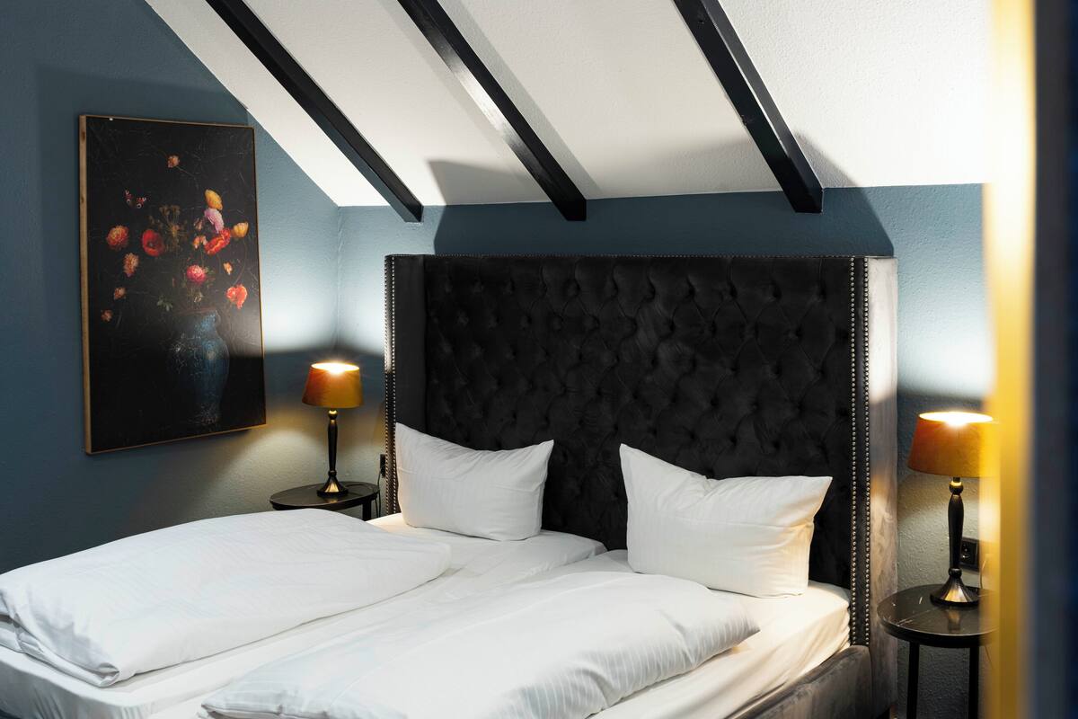 Uma cama bem arrumada com lençóis brancos, duas mesas de cabeceira com luminárias, uma cabeceira escura com detalhes em botão e uma obra de arte emoldurada na parede.