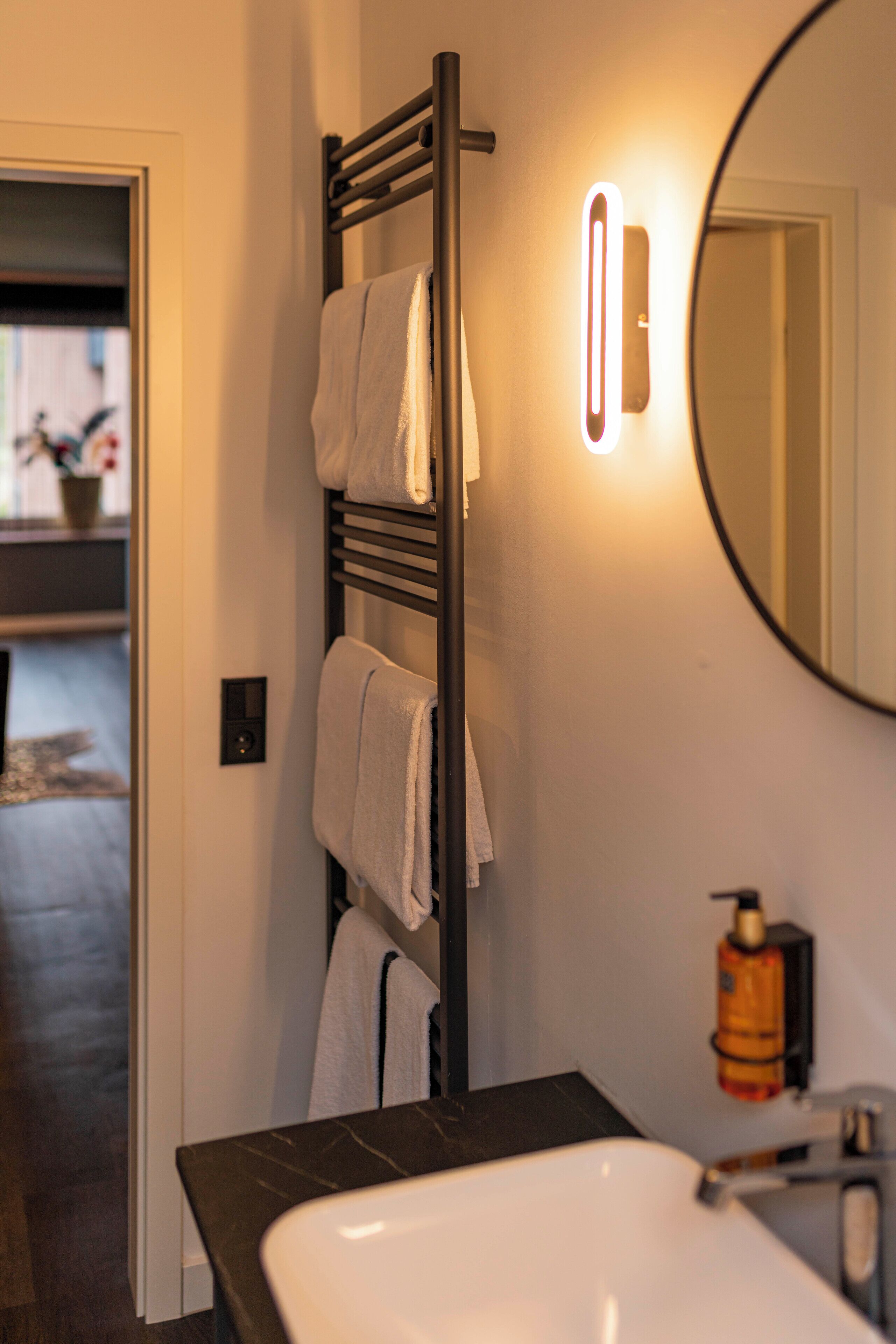 A bathroom with a dark wood towel rack, a white sink, a round mirror, and a wall-mounted light fixture.