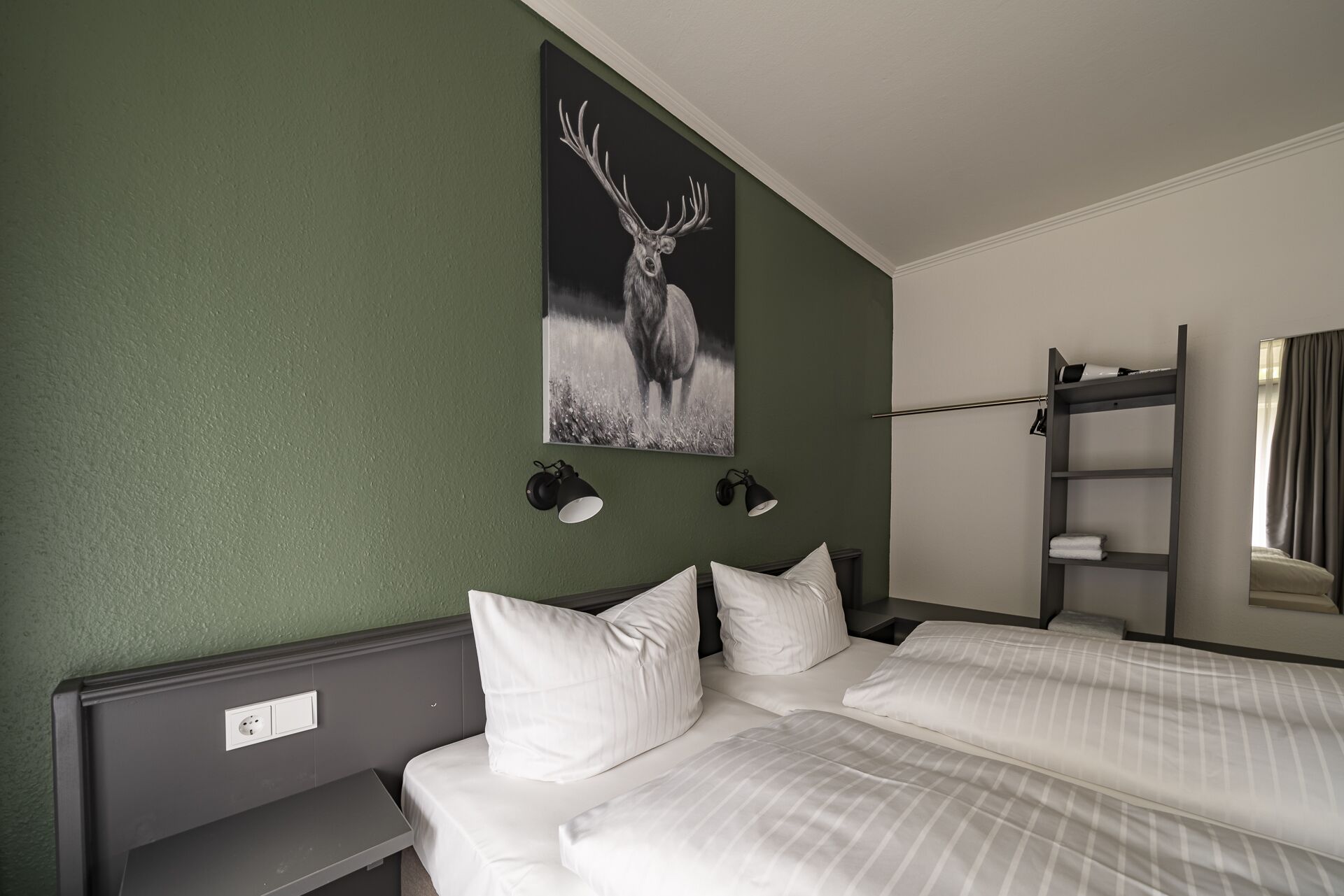 A hotel room with two beds, a green wall, a deer painting, and a shelf with towels.